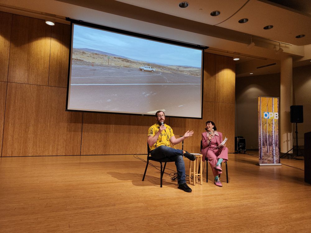 Evan Howington and Lillian Karabaic, seated in chairs on a stage, address the audience