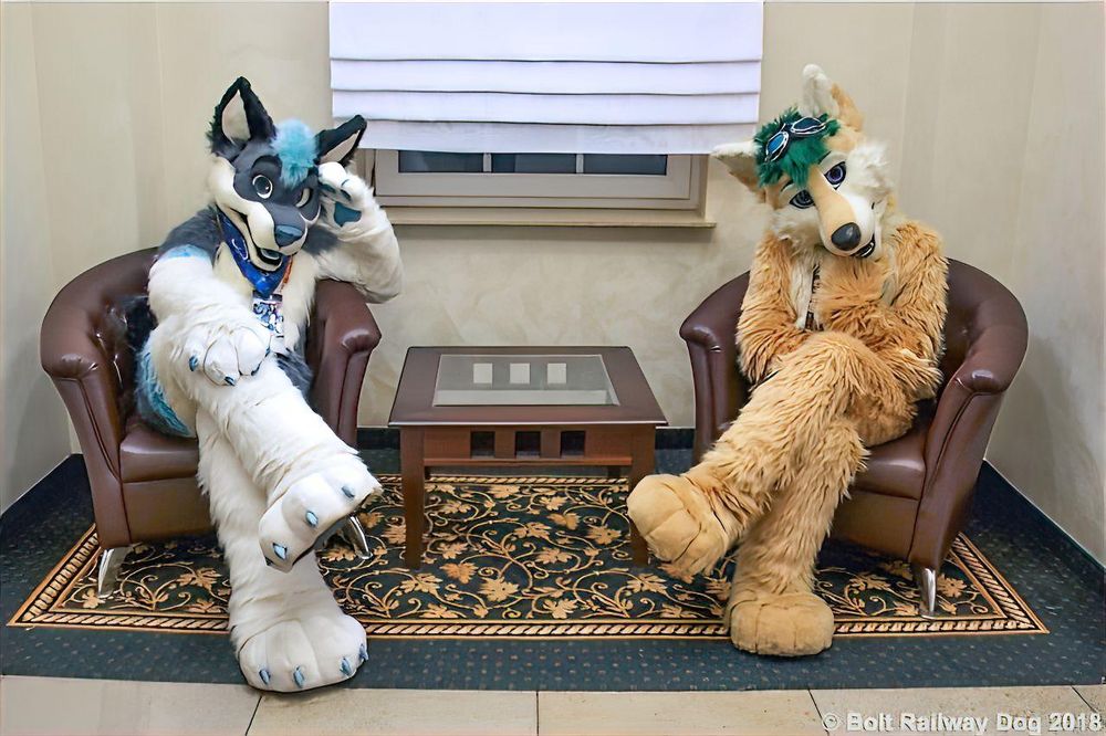 Two husky fursuiters sitting on a chair mimicing the same pose with crossed legs and leaning into their hand on the arm rest. One has beige and blond fur with green hair and the other has Beige and blond husky fursuiter with green hair and purple eyes, the other has grey/blue fur with aquamarine blue marking on forearms, followed by white fur and aquamarine blue claws and hair tuff 
