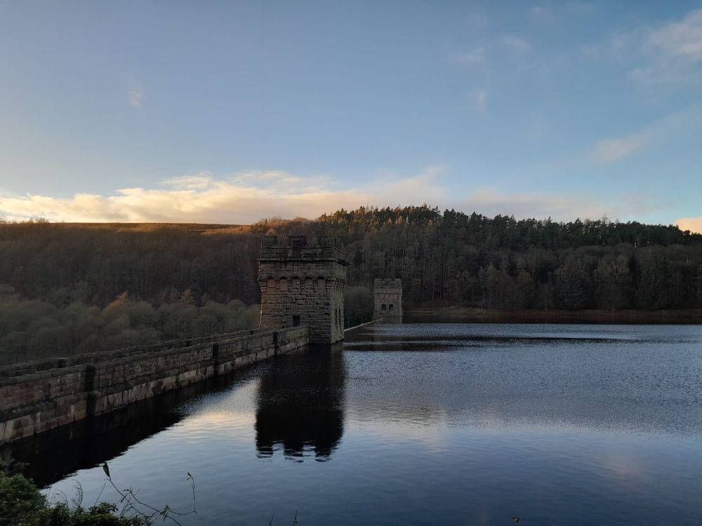 Howden Reservoir. The stone wall and turrets of the dam reflect darkly in still water. Shadowy tree-covered hills stand against the blue sky where clouds still catch the sun.