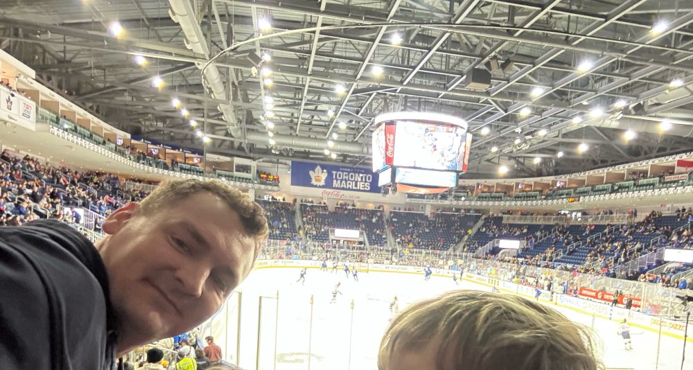 Jeff (middle aged white male with a fresh haircut) sits in the stands at the Toronto Sceptres game. He is smiling at the camera and wearing a blue hoodie as the Sceptres and Frost warm up behind him prior to the start of the game.
