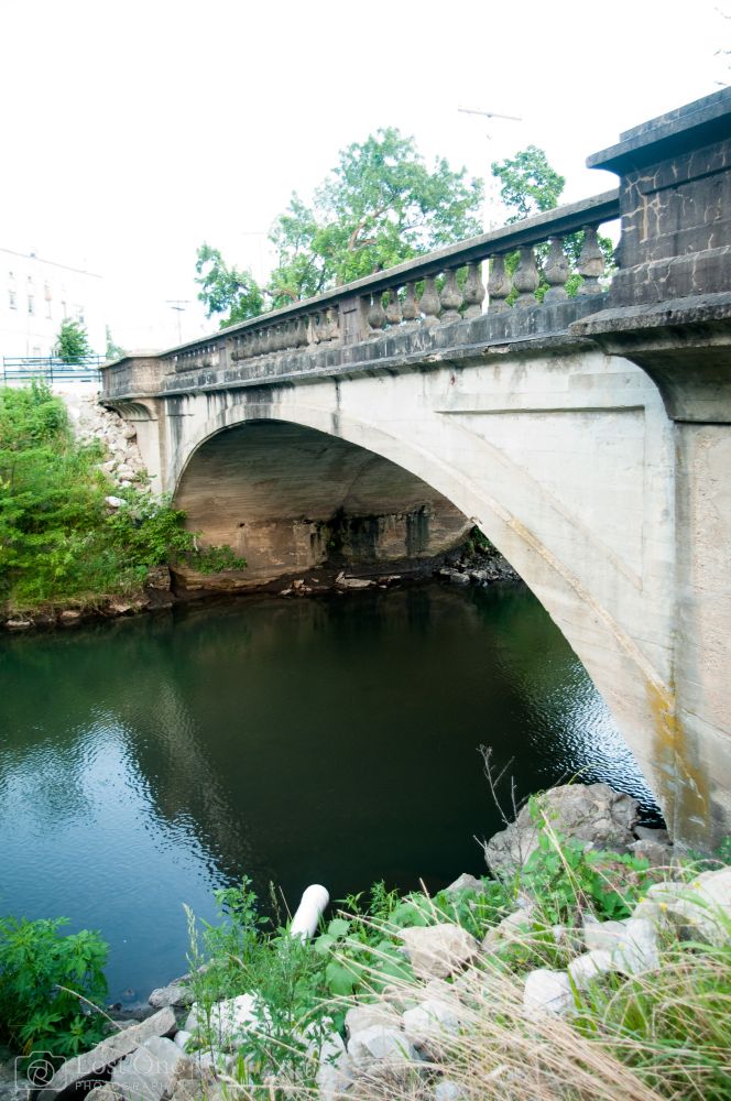 The photo shows an old concrete arch bridge spanning over a calm, dark greenish river. The bridge has a single large arch supporting it, with weathered stonework and streaks of discoloration where water has stained the concrete over time. Above the arch is a balustrade made of short, chunky stone columns lined up in a row, giving it a classic, almost historic look. The railing and edges of the bridge are darkened, either from age, moss, or soot, contrasting with the lighter concrete of the main arch.
The river below is still and reflective, its surface showing ripples of green and blue. The banks around the bridge are overgrown with weeds, shrubs, and patches of grass, with rocks scattered at the edges. On the near side of the river, a large white pipe juts out toward the water, breaking up the otherwise natural look of the shoreline. Jagged rocks and a bit of broken concrete surround it. On the far bank, the base of the bridge has exposed stone and concrete, with vegetation creeping along its edges.
In the background, the top of the bridge leads into a tree-lined street. The trees are lush, green, and partially obscure what looks like a pale stone or brick building to the left. Power lines cut faintly across the sky, which is washed out and very bright, almost overexposed, making the bridge and greenery the strongest visible features.
The overall mood is one of quiet age and utility — a small town or older city bridge that has stood for decades, carrying traffic over a modest river. The scene feels still and slightly worn, but also rooted, like it belongs exactly where it is.