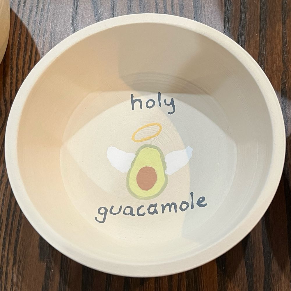 A small in progress ceramic dip bowl for guacamole. It has a drawing of an avocado with a halo and wings and says “holy guacamole”