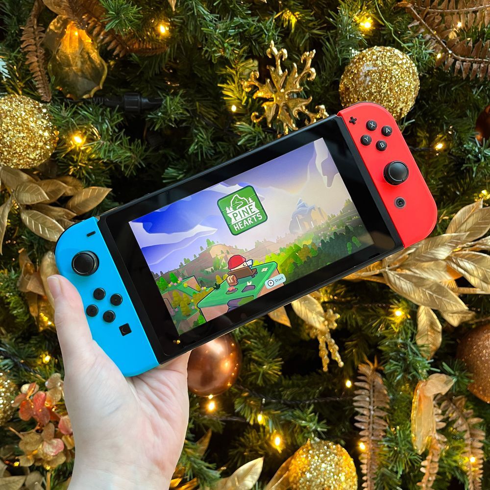 A hand holds up a Nintendo Switch in front of a Christmas Tree. Pine Hearts is running on the Switch.