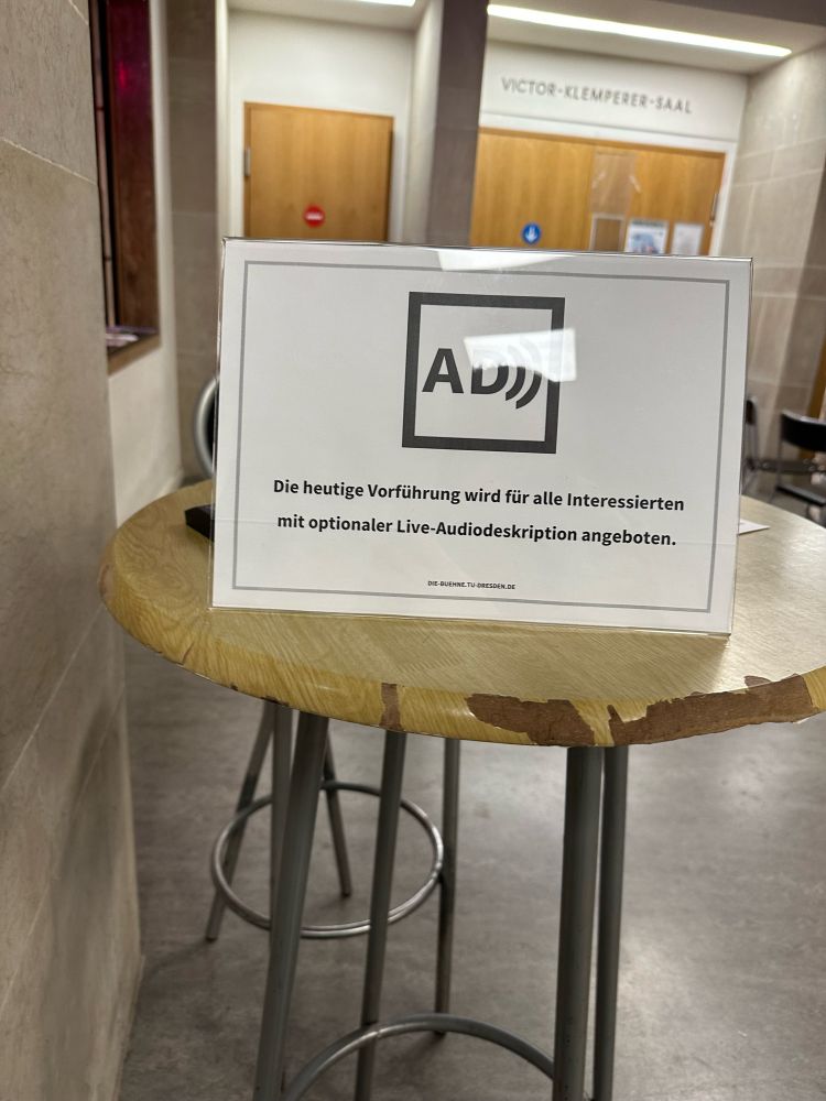 Auf einem Bartisch steht ein Hinweisschild mit Symbol für Audiodeskription. Dazu der Text: „Die heutige Vorführung wird für alle Interessierten mit optionaler Live-Ausiodeskription angeboten.