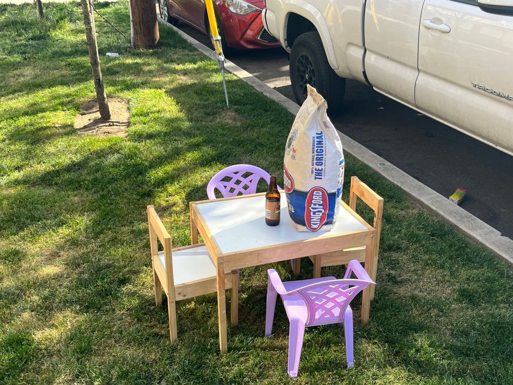 a child sized table with mismatched chairs is on a grassy curb. placed on top of the table is a full bag of concrete and an opened bottle of root beer