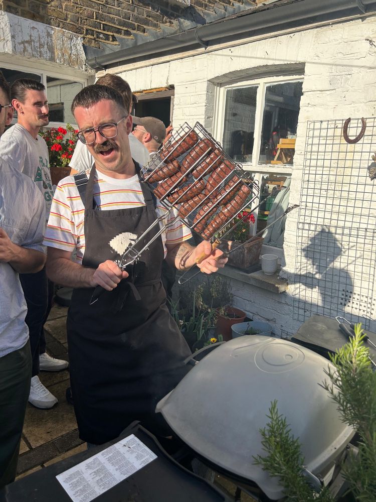 Man in a black apron holding a barbecue cage filled with beef sausages. He’s standing over a gas barbecue that is grey and whites, with a rosemary bush in the foreground behind him a white wall and a window and some men