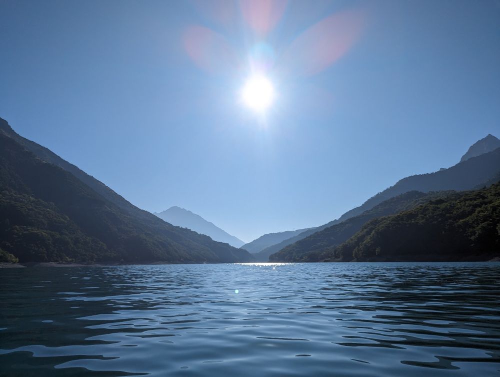 A scenic view of mountains and a lake. Camera is on a boat close to the water. The sun is in the middle of the image between two mountains.