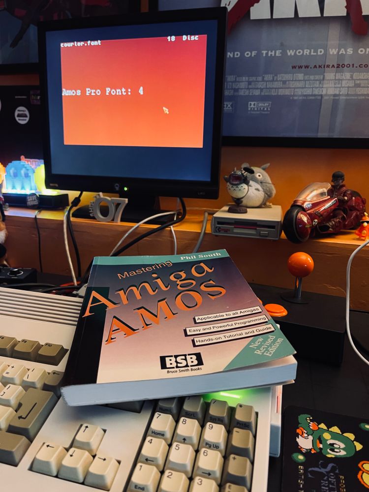 The book 'Mastering Amiga Amos' rests on top of an Amiga A1200 computer. A monitor in the background has some font information on the screen.