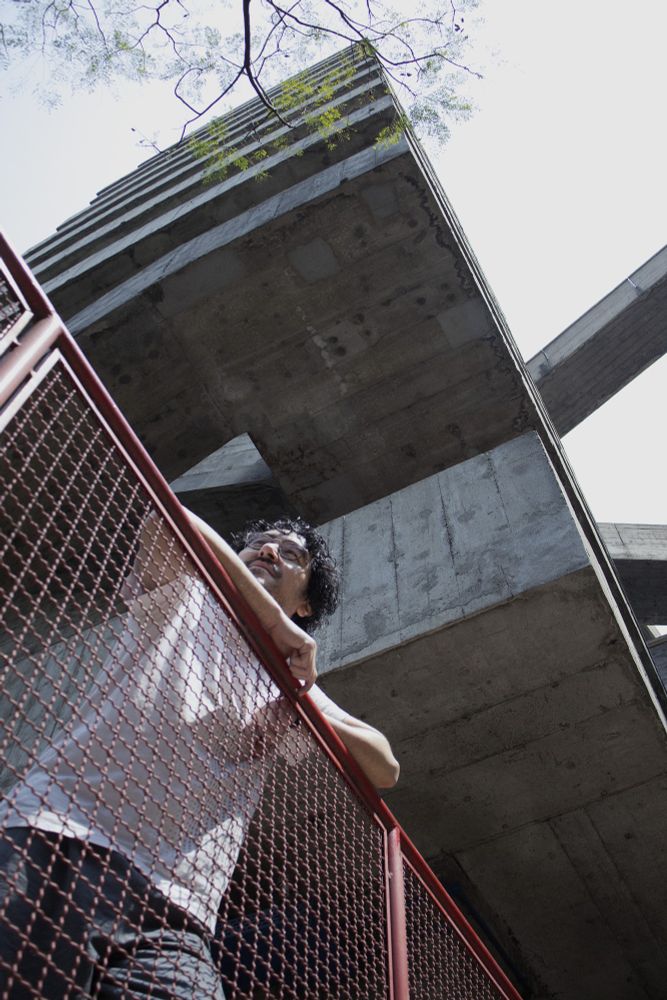 Foto tirada de baixo para cima de um homem com traços orientais usando óculos, camiseta branca e calça cinza. Está apoiado em uma grade vermelha e ao fundo um prédio com concreto aparente. Foto tirada no SESC Pompéia.