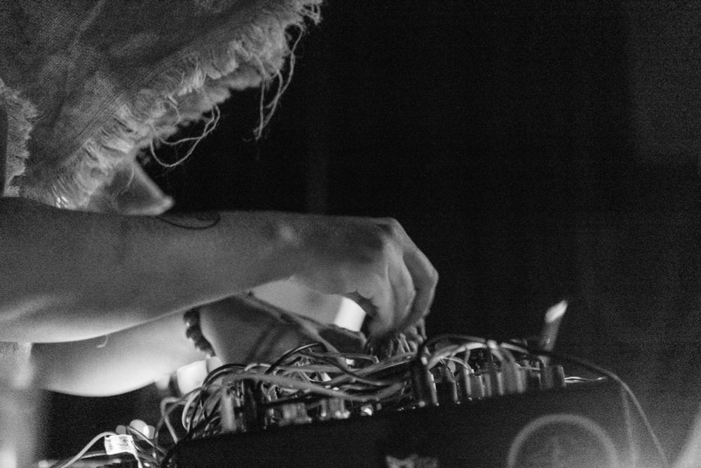 Close up of the burlap hooded man's hands, performing precise adjustments to a tangled nest of cables plugged into the arcane synth.