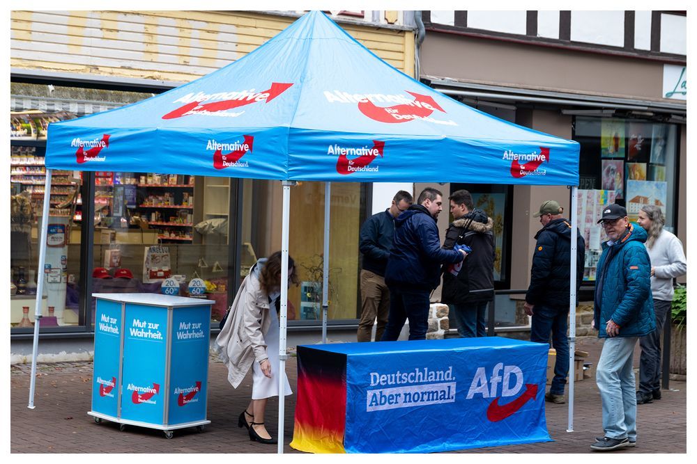 AfD Infostand mit Pallivion und Infotischen anwesend und auf dem Bild zu sehen mehrere Aktive im Kreisverband Göttingen vor einer Häuserzeile. Um das Bild ist ein kleiner weißer Rahmen.