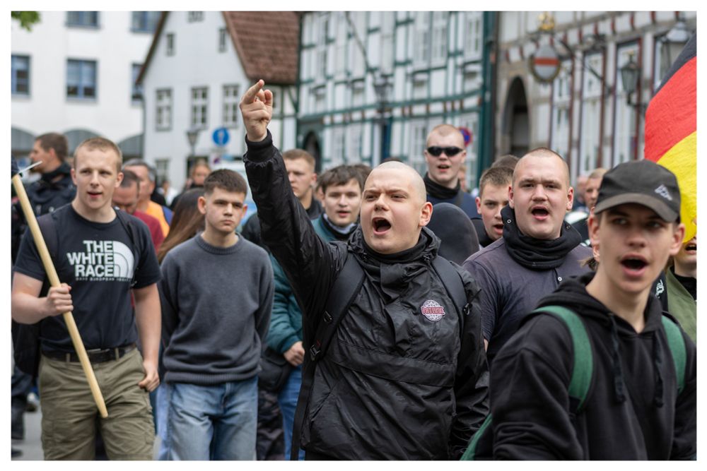 mehrere Neonazis auf dem Bild. Der Neonazi in der Mitte streckt einen Mittelfinder in die Höhe.
Um das Bild ist ein kleiner weißer Rahmen