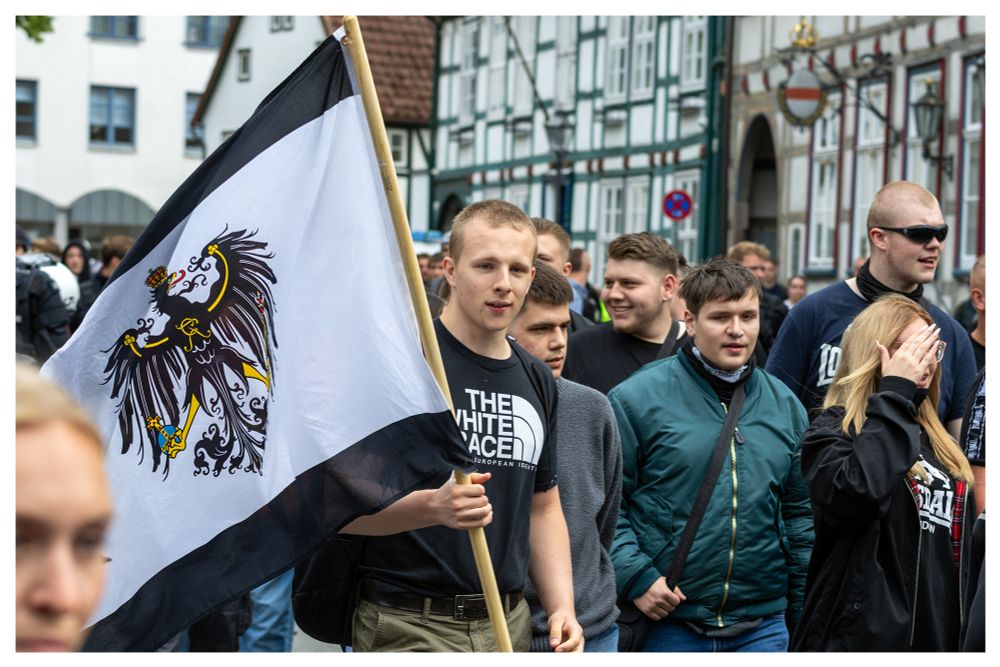 Mehrere Neonazis auf dem Bild zu sehen. Ein Neonazi mit einem "THE WITHE RACE" Shirt trägt eine alte Preußen Fahne. 