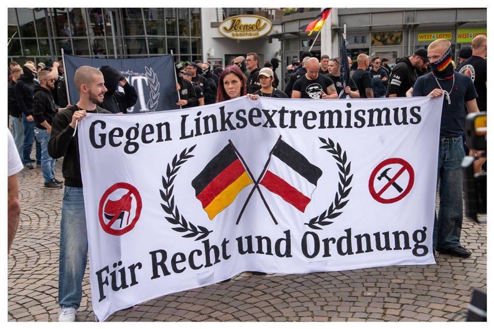 Neonazis hinter einem Transparent mit der Aufschrift:" Gegen Linksextremismus - Für Recht und Ordnung" Zwischen dem Spruch gekreuzt in einem Lorbeerkranz eine  schwarz-weiß-rote und schwarz-rot-gelbe Fahne. Neben dem Lorbeerkranz die Antifa Fahnen und ein Hammersymbol, beides im Stiele eines Verbotsschid durchkreutzt. Im Hintergrund mehrere Teilnehmende des Neonazi Aufzugs.
Um das Bild ist ein kleiner weißer Rahmen
