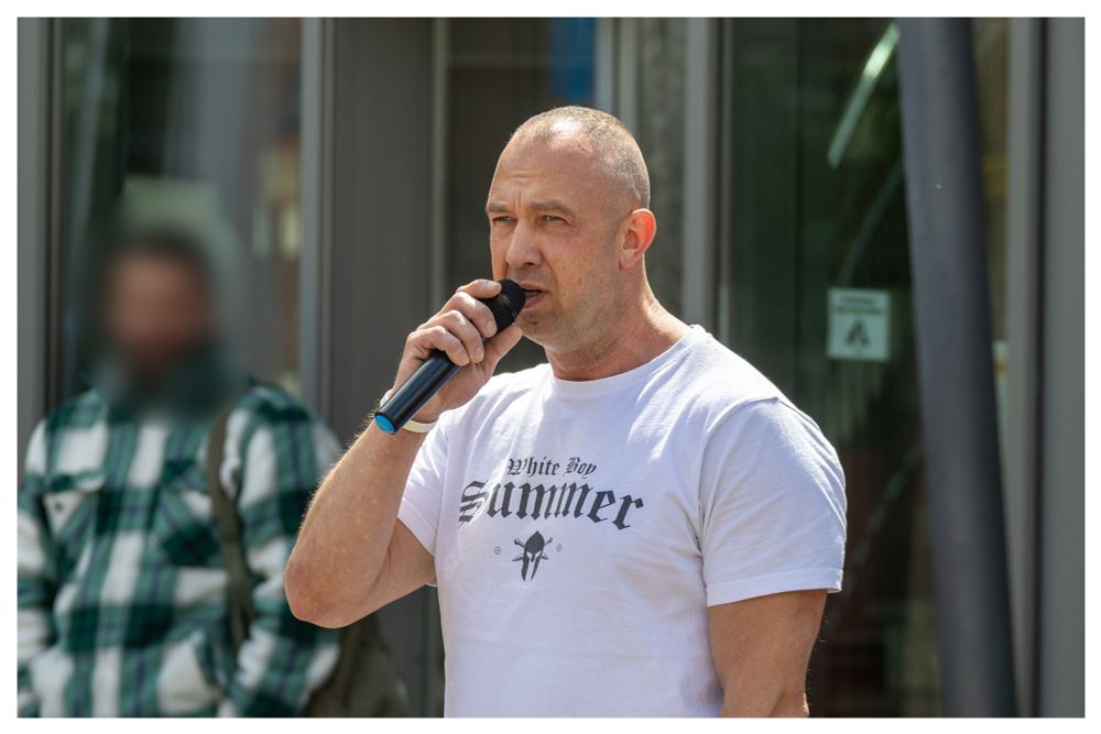 Neonazi Daniel Kokott, ein stämmiger Mann mit Glatze und weißem "White Boy Summer" Shirt spricht in ein Mikrofon.
Um das Bild ist ein kleiner weißer Rahmen