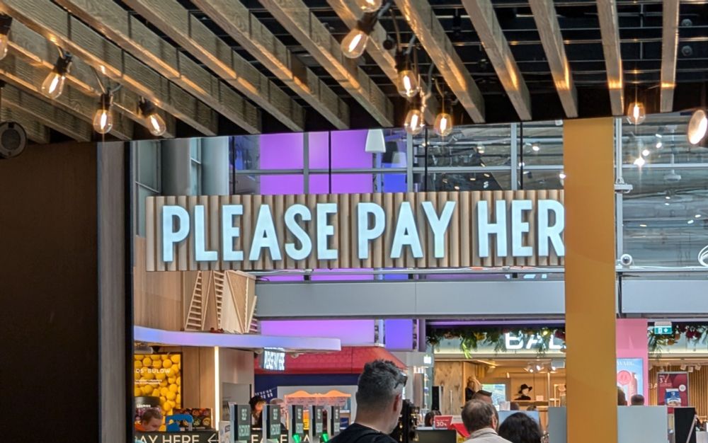 A duty free cash register with "PLEASE PAY HERE" in large letters over it, but the last E is obscured by a pillar