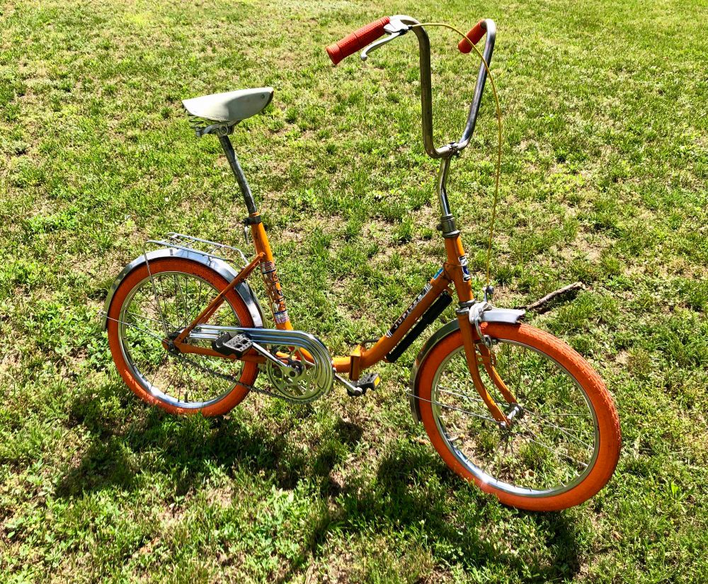 Ein frisch restauriertes orangenes Klapprad aus dem 80er Jahren steht auf einer Wiese, die Chromfelgen glänzen in der Sonne