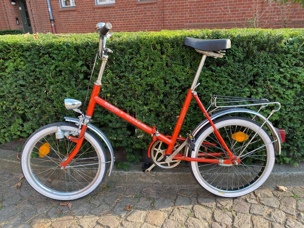 Ein hellrotes Mifa-Klapprad vor einer Hecke