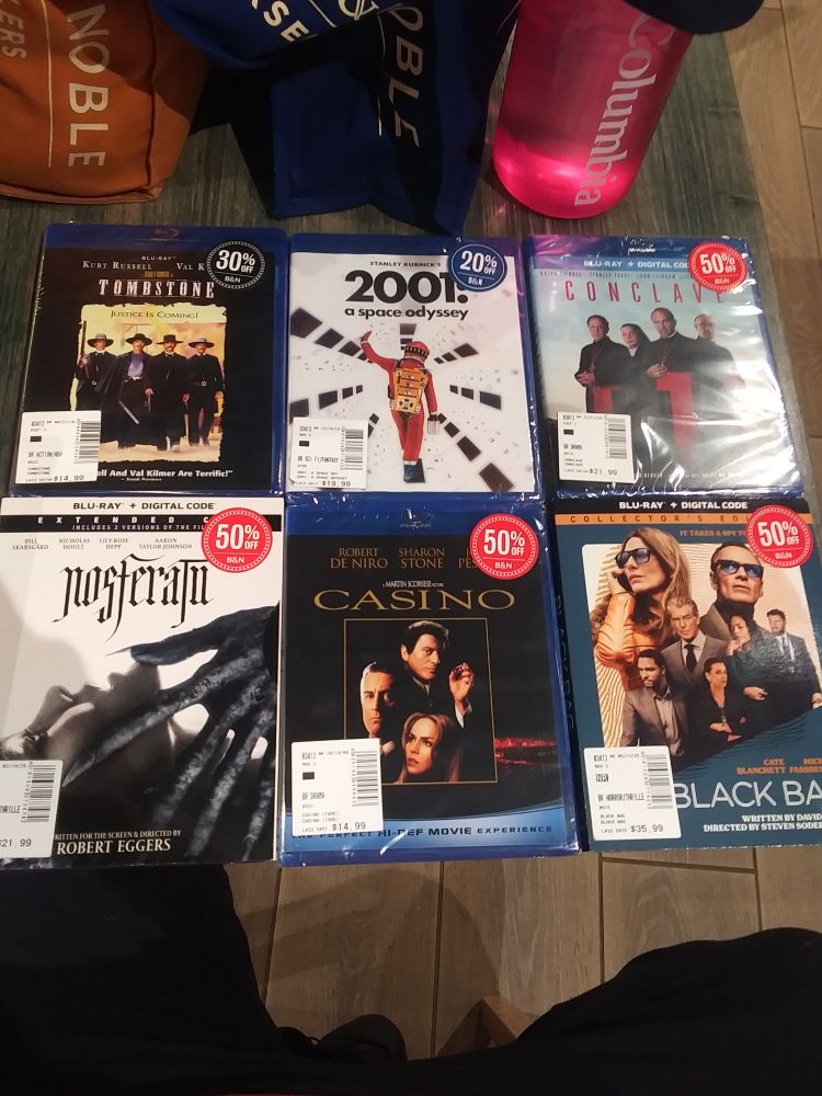 Six movies are shown: Tombstone, a nineties western with four men on the cover in black jackets and black cowboy hats
2001 A Space Odessey: A man in an orange space suit is walking down a white bright corridor of a space ship.
Conclave: Four clergymen are on the cover looking straight ahead. 
Nosferatu: Close up of a woman looking up in the air while the spindly hands of a vampire touch her chin.
Casino: Two men in business suits and a woman with long brown hair are on the cover.
Black Bag: Seven people are on the cover. All are wearing nice clothes, suit and ties. And two of them have black sunglasses on. 