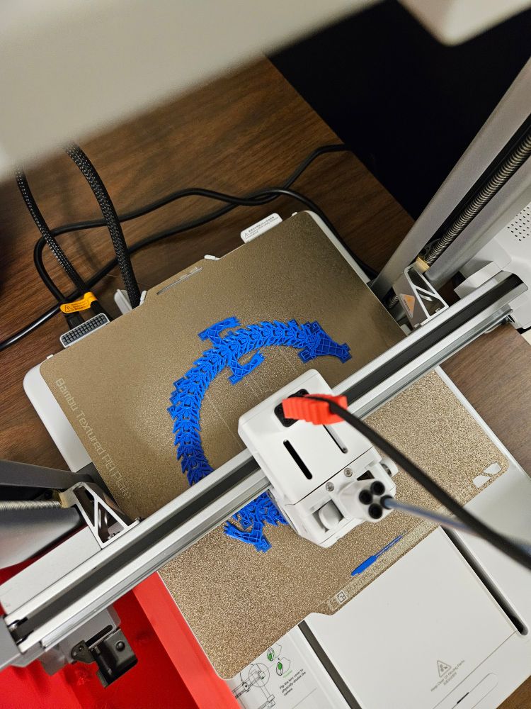 There is a Bambu A1 printer, with a "birds-eye-view" or a dragon that is in progress. The links are thin and overlapping, and the feet are around 20 layers thick. 