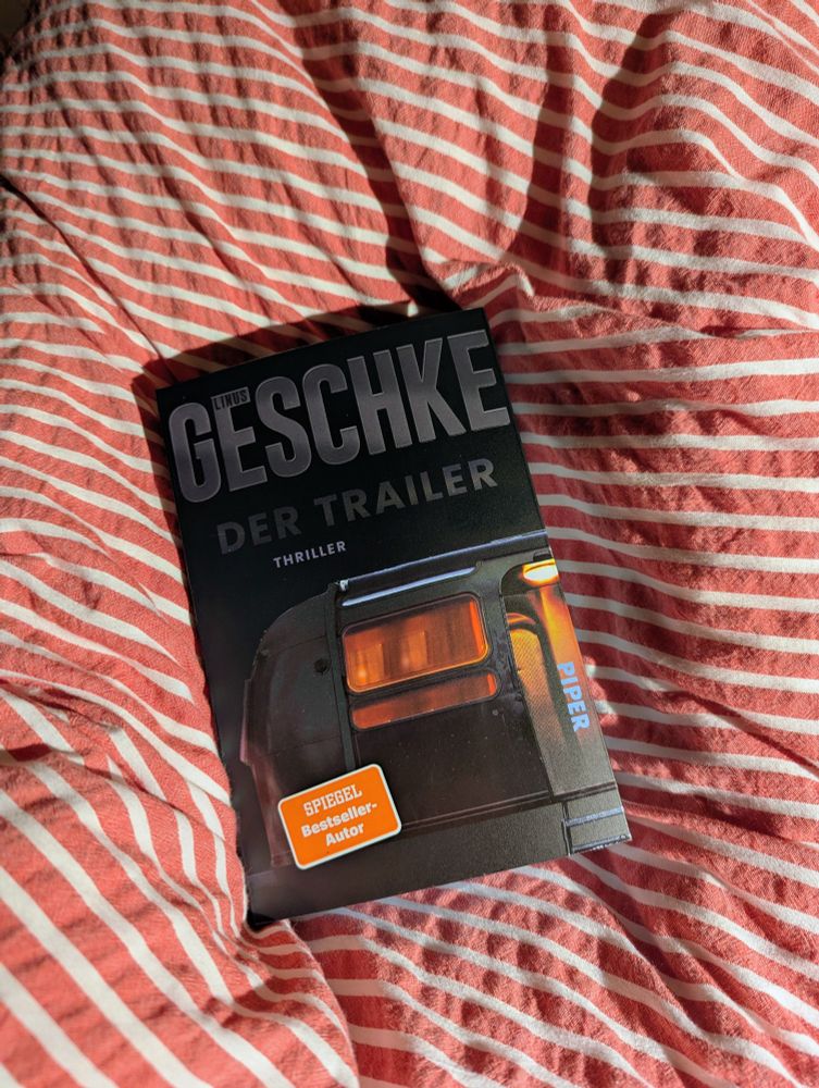Das Bild zeigt ein Buch mit dem Titel Der Trailer" von Linus Geschke, einem Spiegel-Bestseller-Autor, das auf einem rot-weiß gestreiften Stoff liegt.  Das Cover des Buches ist schwarz und zeigt ein Bild eines Wohnwagens mit beleuchteten Fenstern.
