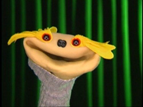 A smiling tan sock puppet in a grey turtleneck with a small black doggy nose and large yellow petals around its red rimmed eyes