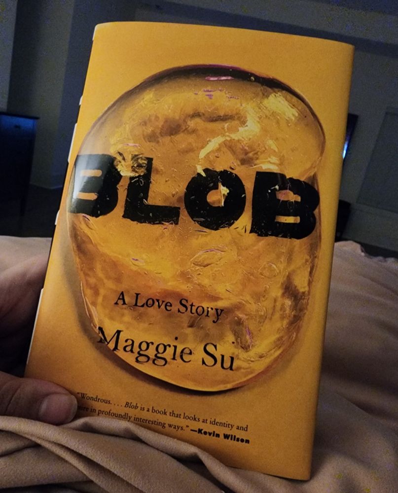Me, a white lady, is holding up a copy of the book Blob by Maggie Su. It has a wonderful yellow cover with black font, as seen through a gelatine blob. 