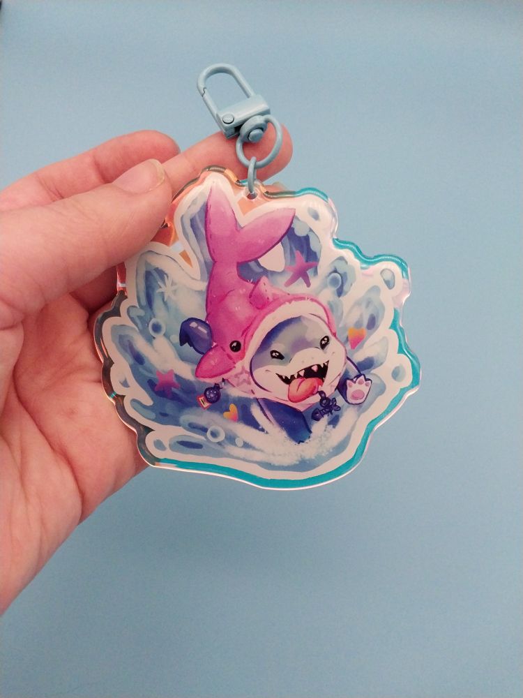 A rainbow acrylic keychain featuring Jeff the Shark wearing a pink dolphin costume, running happily with splashing water behind him.