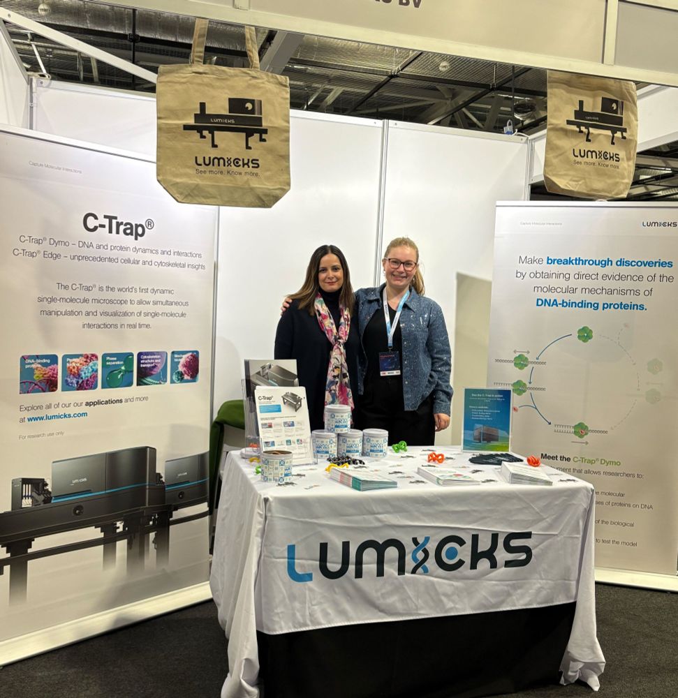 Two persons welcoming UK biophysicists behind a table laden with educational and marketing materials on dynamic single molecule analysis and the C-Trap.