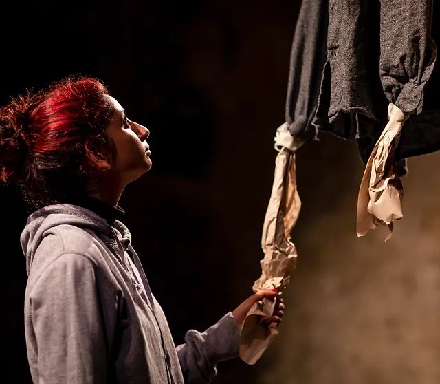 Don’t Smoke in Bed Production photo. A young woman in grey hoodie holding onto the leg of a paper creature hanging from the sky. 