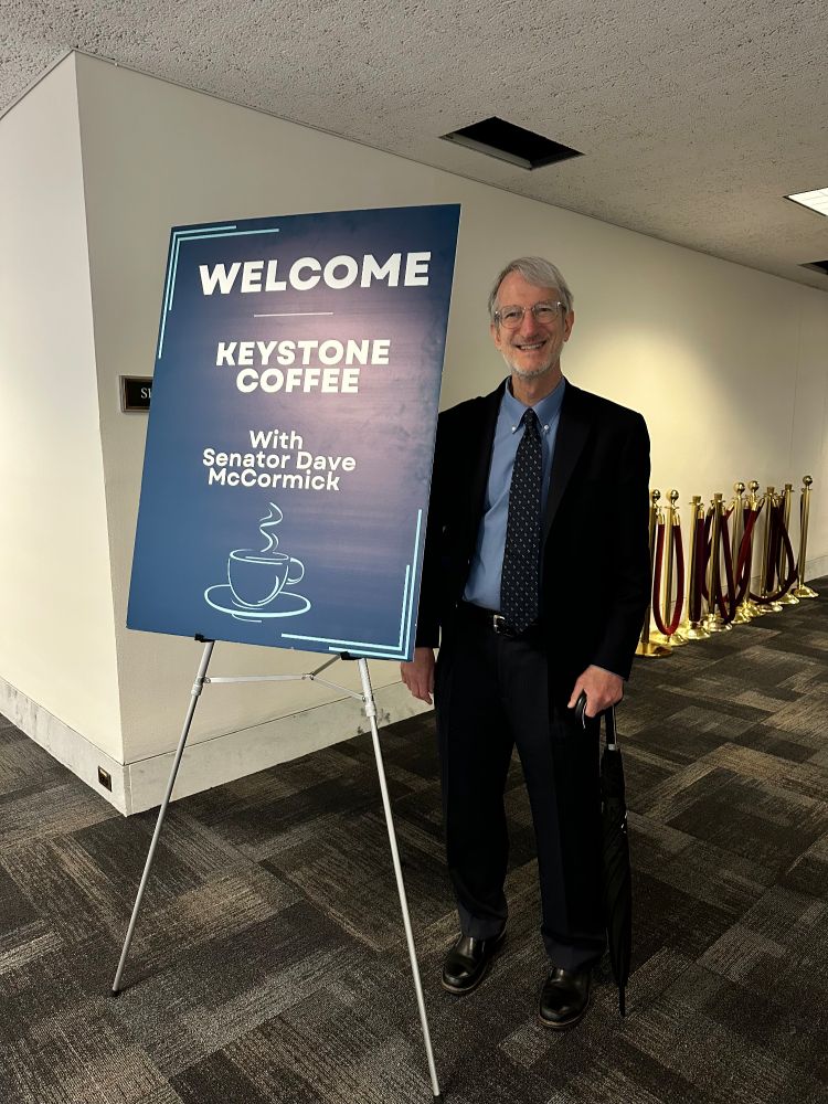 An ASBMB members smiles while standing next to a sign that reads Welcome. Keystone coffee with Senator Dave McCormick. 