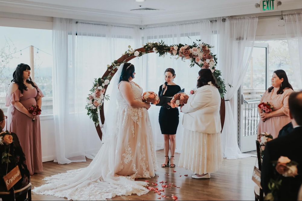 Shannon and Janette’s wedding. They are in front of the officiant and holding their bouquets as their bridal party stands to the side 