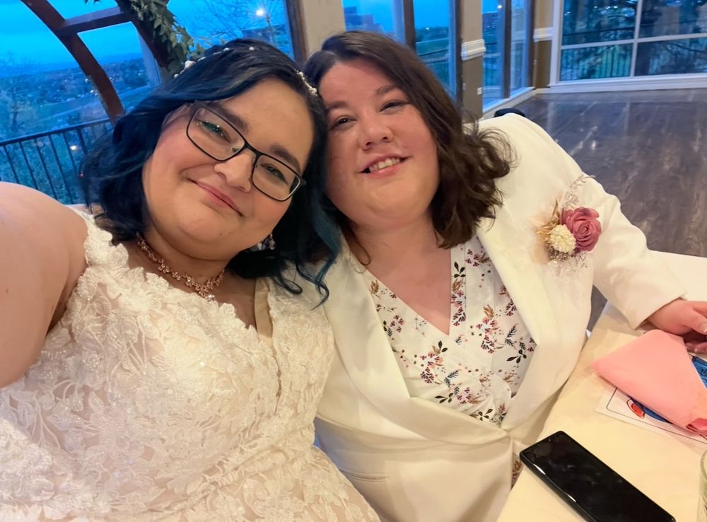 Shannon and their wife Janette taking a selfie at their wedding at the bridal table during the reception. Shannon has blue hair and a wedding dress with spaghetti straps. Janette has brown hair, a white blazer with a floral white shirt 