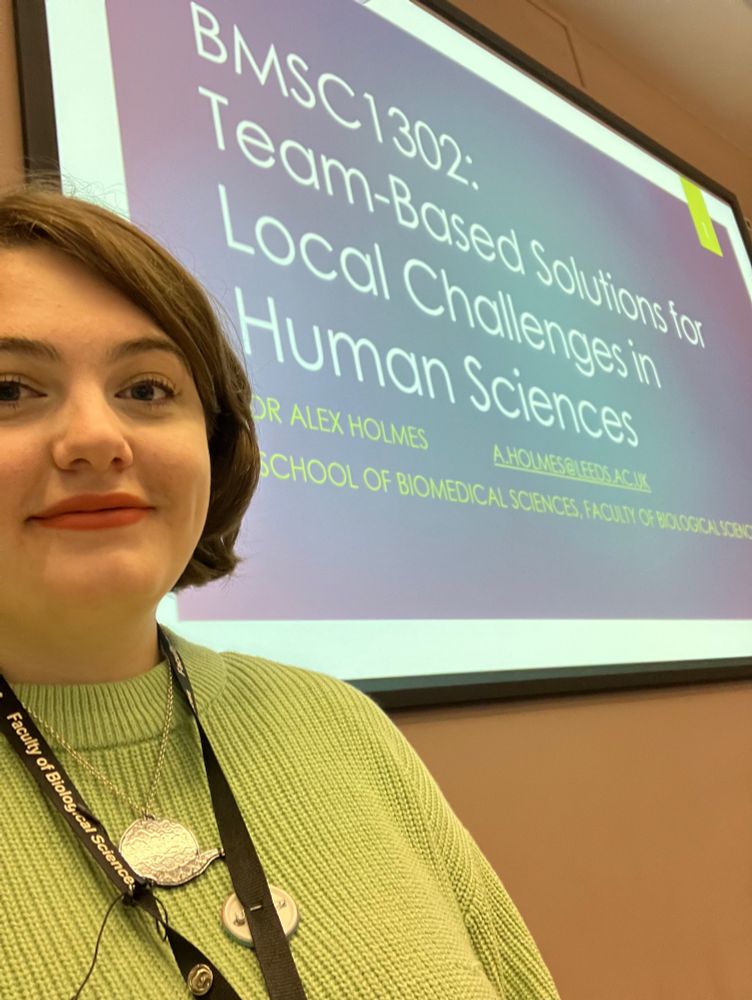 Alex wearing a green jumper and smiling in front of a large screen reading “team
based solutions for local challenges in human sciences”
