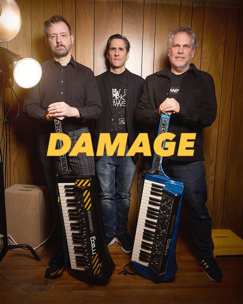 Press photo of the band Damage. Photo depicts three people, the two in the foreground holding analog synthesizers.