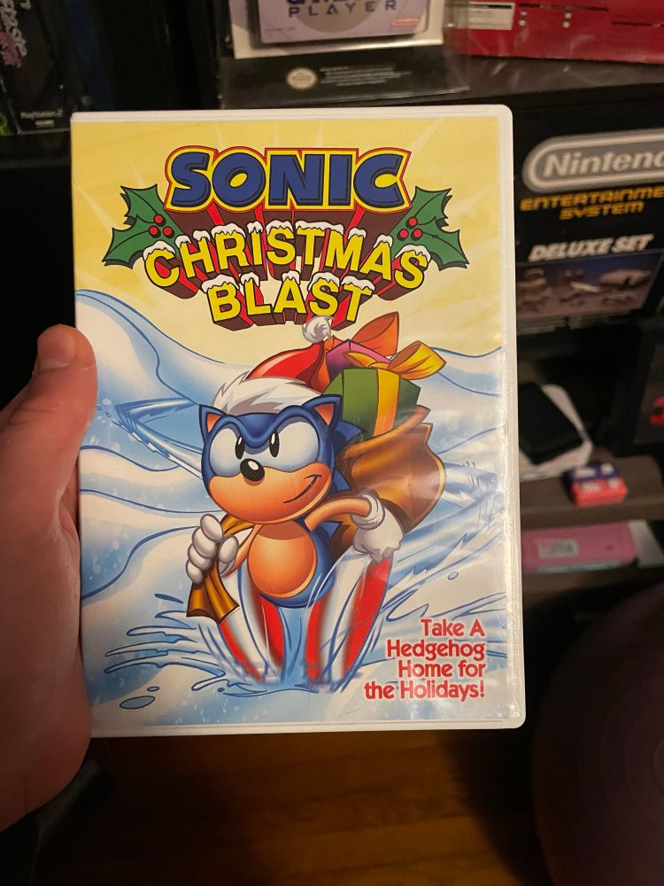 Front cover of Sonic Christmas Blast DVD with Sonic peeling out in the snow wearing a Santa hat and bearing presents