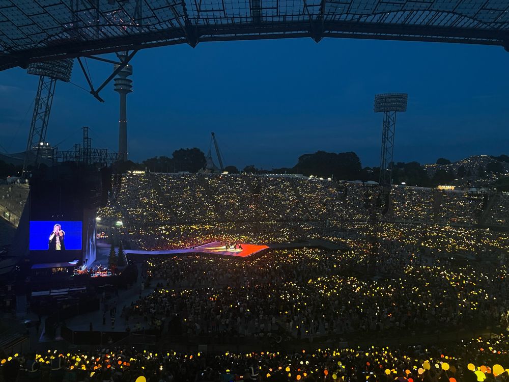 A picture taken from the nosebleeds at eras tour Munich. It shows the whole crowd, holding up luminous orbs in yellow and orange during willow. We see a screen on the left side of the image displaying Taylor pointing at us while singing. In the background we can see a hill outside the stadium that is filled to the brim with swifties, who are also holding up orbs or flashlights 
