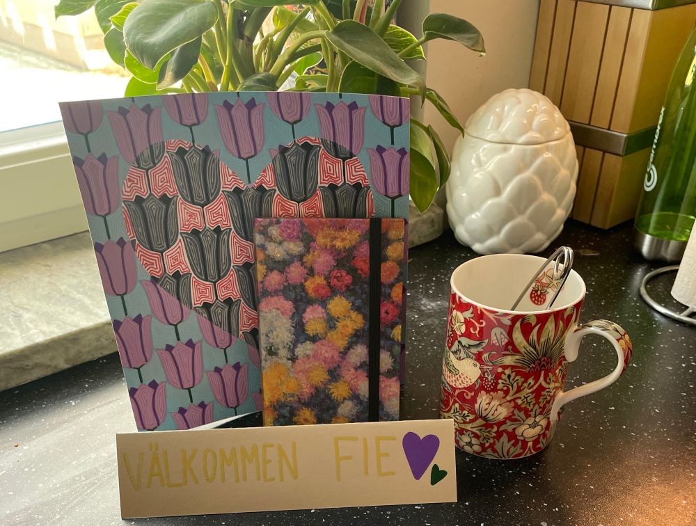 A setup on the kitchen table of cute things from my girlfriend. Including a mug with favourite tea prepared, a little flowery notebook, a “Välkommen Fie” (welcome Fie) sign with hearts on it, andddd a painting. 
The painting looks like a print of purple tulips on a turquoise background, and in the shape of a heart, the background is changed to red and the tulips to grey. 
- tulips are one of my favourite flowers, and purple is one of my favourite colours