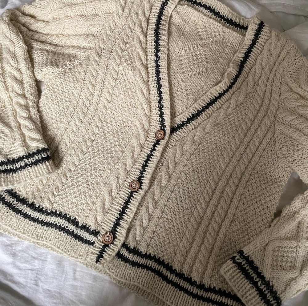 A hand knit cardigan looking very similar to the original folklore one. It’s cream with dark grey stripes in the ribbing, and with wooden buttons