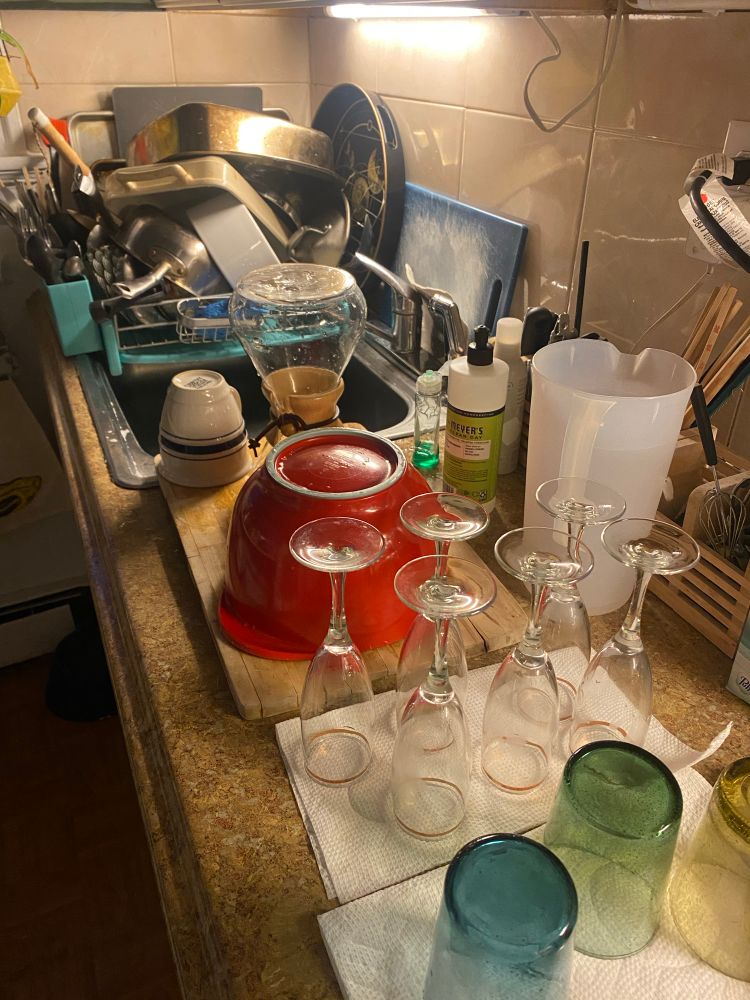 A drying wrack and counter top covered with drying glasses, pots, pans, mugs, cutlery, and champagne flutes. 