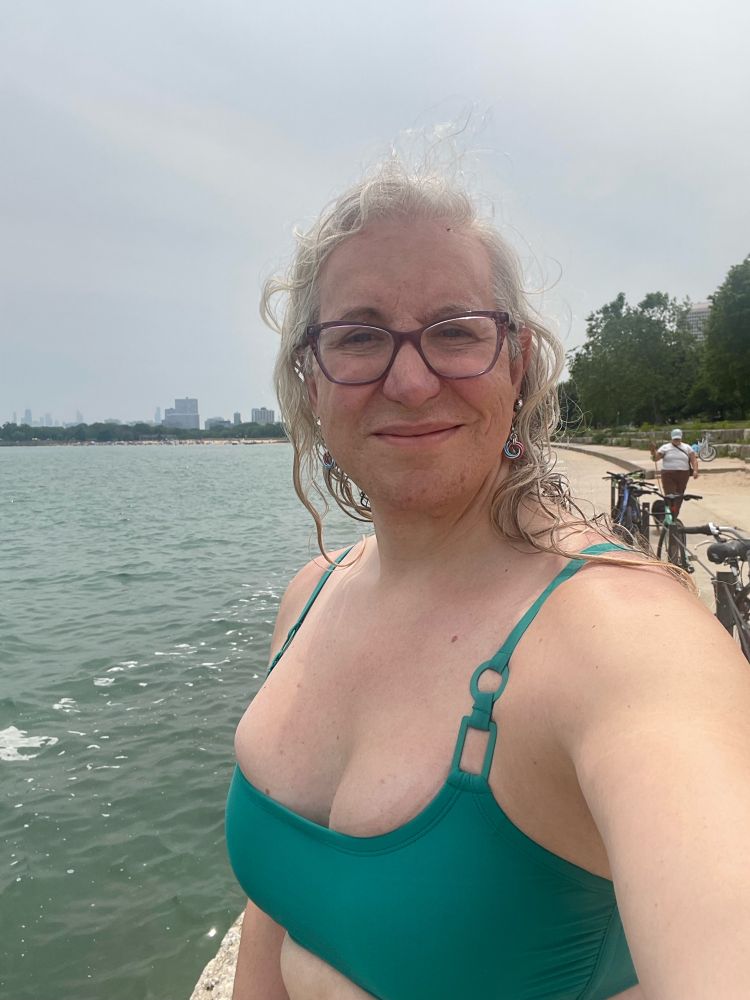 A gal taking a selfie, in a teal bikini. Behind her is the water and a city skyline.