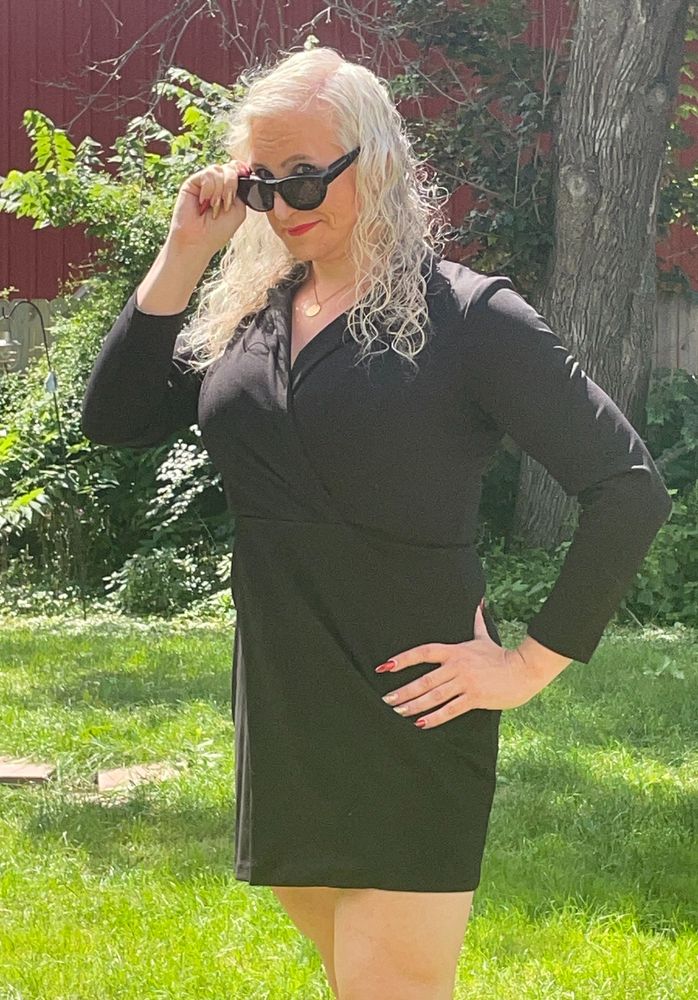 A gal dressed in a black dress, long sleeves on top and shorter skirt on bottom. She's looking at the camera but has pulled her sunglasses down. Her other hand is on her hip. Some of her nails are red, others are glittery gold. She has long white curly hair. She is outside on a sunny day. 
