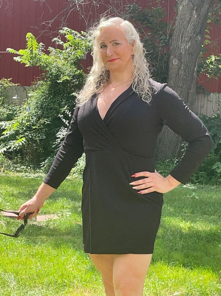 A gal dressed in a black dress, long sleeves on top and shorter skirt on bottom. Her sunglasses are being held in one outstretched arm, her other arm is on her hip with her elbow out. She's looking at the camera contemplatively. She has long white curly hair. She is also outside on a sunny day.