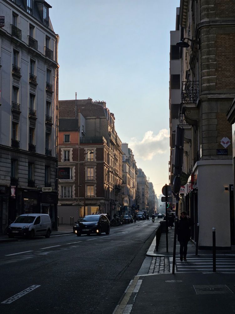 Rue parisienne bordée d’immeubles sous une lumière douce, avec quelques voitures et passants en silhouette sur le trottoir, et un ciel clair en arrière-plan.