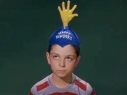 A white boy of about 10 years old wearing a red and grey striped shirt and a blue beanie. The blue beanie has a yellow hand sticking out of the top, and white lettering that reads HAPPY FINGERS.