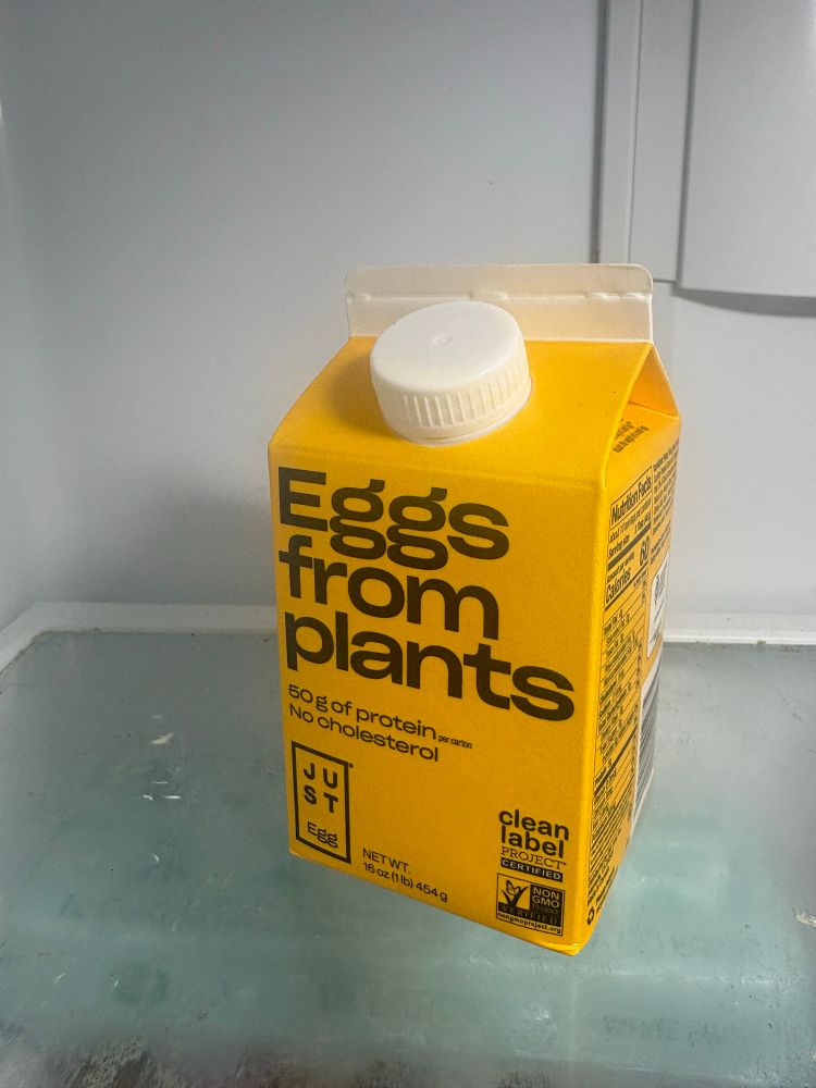 Closeup photo of a bright yellow and white carton sitting inside a refrigerator. The carton has black text on it that reads “eggs from plants” as it is a carton of plant-based egg substitute. Below the frosted glass shelf that the carton is sitting on you can see a carton of eggs.
