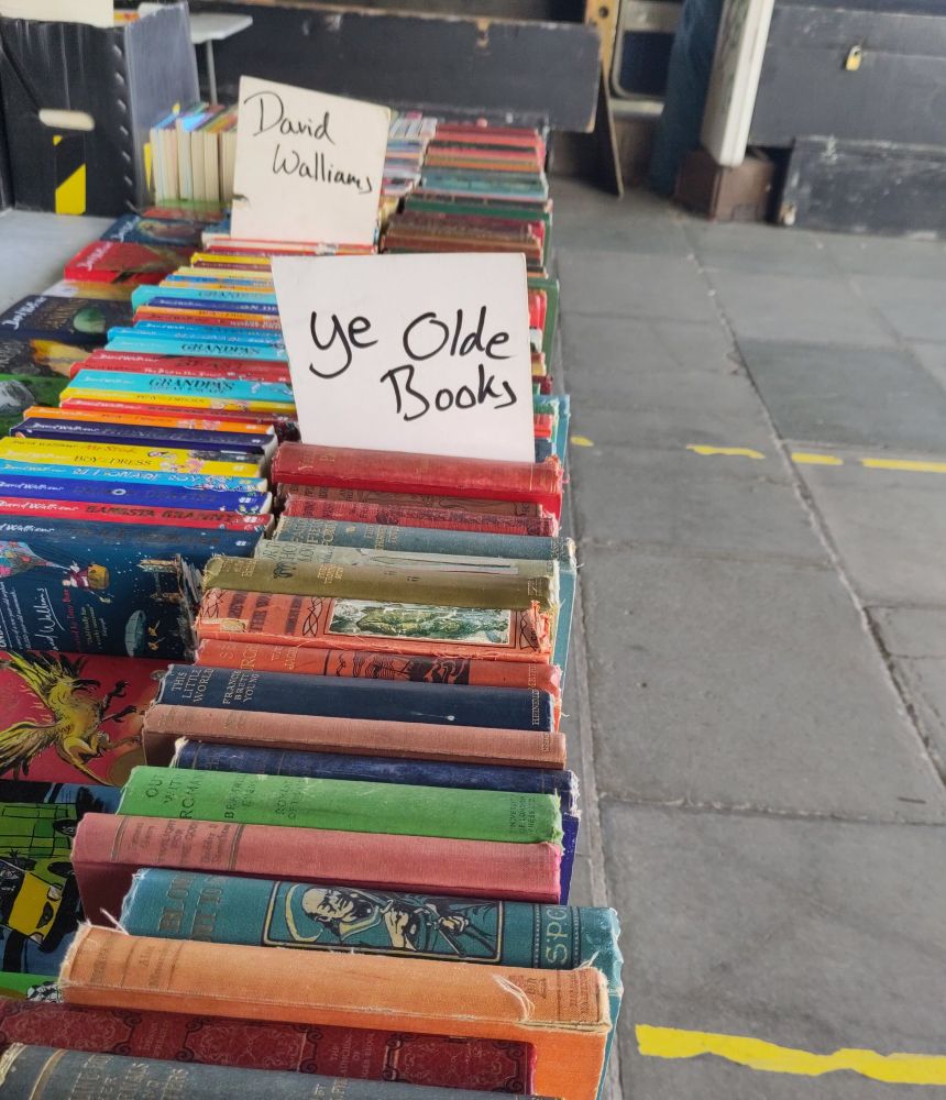 Two rows of books at a street bookstall. There are two handwritten signs on dividers. One says "Ye Olde Books", the other says "David Walliams" 