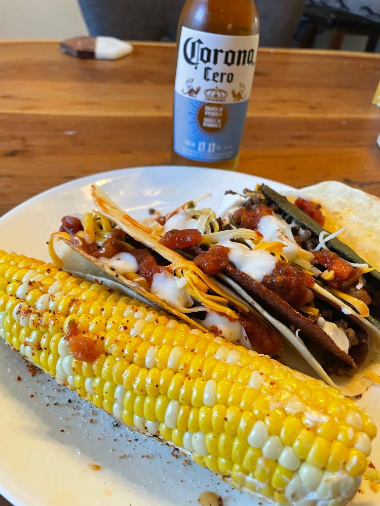 3 hamburg tacos, one with a green (jalapeño) hard shell, one red (chili), & one yellow corn hard shell, wrapped with soft wheat tortillas. Corn on the cob sits on the side of the plate. Non alcoholic Corona behind. 