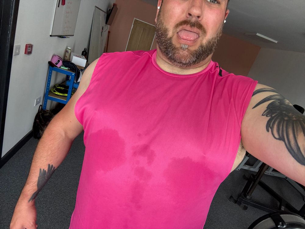 A person is shown wearing a pink sleeveless shirt with sweat marks, sticking out their tongue. The background features gym equipment and a blue shelf.