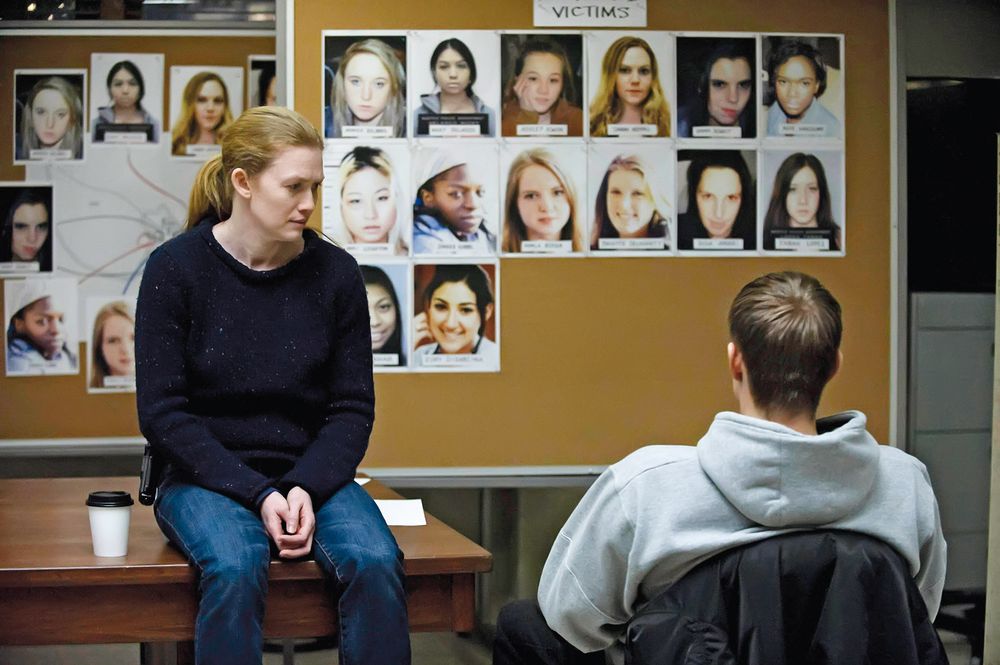 Detective in navy sweater sitting on a desk talking to a suspect in a grey hoodie in front of a police investigation board covered with photos of female victims, labelled "Victims," in a crime investigation room.